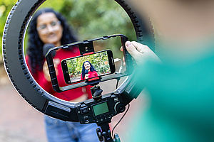 Auf dem Foto sieht man eine Person mit Handykamera und Ringlicht, die eine Frau mit Mikrofon filmt..