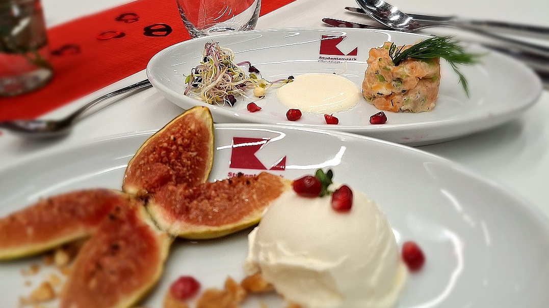 Alt-Text: Ein festlich gedeckter Tisch in einer Mensa für ein Valentinsdinner, auf dem zwei weiße Teller mit elegant angerichteten Speisen im Fokus stehen. Im Vordergrund zeigt ein Teller ein Dessert: eine Kugel helles Eis (oder Cremespeise), garniert mit Granatapfelkernen, daneben aufgeschnittene frische Feigen und gehackte Nüsse. Im Hintergrund steht ein weiterer Teller mit einer Vorspeise, bestehend aus einem Tatar (Lachs), Sprossen, einem Klecks heller Soße und Granatapfelkernen. Die Teller tragen ein rotes Logo des Studierendenwerks. Dahinter stehen ein Wasserglas mit einer Schleife und ein roter Tischläufer. Im unscharfen Hintergrund ist ein Schild mit der Aufschrift „Tisch 3“ zu erkennen.