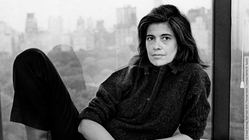 Susan Sontag posiert auf Fensterrahmen liegend. Sie trägt eine dunkle Hose und einen Pullover. Das Bild ist schwarz weiß und im Hintergrund ist die New Yorker Skyline zu erkennen.