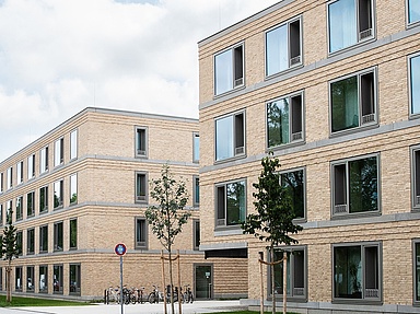 Wohnheim Chiemgaustraße, Neubauten Auf dem Foto sieht man zwei Wohnblocks in einer Straße. Es sind Neubauten mit Flachdach, hellbraun geklinkert, bodentiefe breite Fenster. Vor den Häusern ein kleiner Grünstreifen mit jungen Bäumchen.