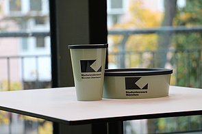 Die StuBowl hat Gesellschaft bekommen in Form des StuCup. Das Bild zeigt ein StuCup neben einer StuBowl (jeweils mit STWM-Logo versehen) auf einem Mensa-Tisch. im Hintegrund ist ein Fenster mit Blick ins Grüne.