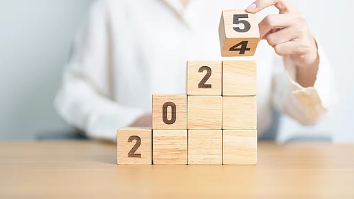 Eine Person im weißen Hemd sitzt an einem Holztisch und stapelt Holzklötze wie eine Treppe auf. Die Klötze zeigen die Jahreszahlen beim Übergang von 2024 zu 2025. Die Zahlen „2“, „0“ und „2“ bilden die ersten Stufen, während die Hand der Person gerade den obersten Klotz dreht, um die Zahl „4“ durch die „5“ zu ersetzen.
