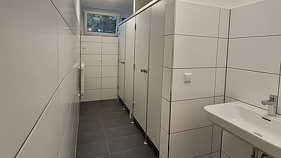 Sanierte-Duschen-Studentenstadt-Freimann-Studierendenwerk Raum mit Duschkabinen und Waschbecken - sieht unbenutzt aus, sehr neu und sauber, weiße, große Fliesen an den Wänden, neue Kabinentüren, neuer Fußboden.