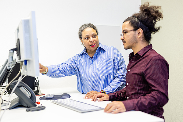 Zwei Personen stehen an Büroarbeitsplatz. Auf dem Foto sieht man zwei Kollegen/-innen, die an einem Büroarbeitsplatz mit PC stehen. Die eine Person zeigt auf den Monitor und erklärt der anderen etwas.