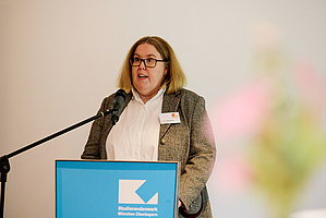 Auf dem Foto sieht man eine Frau an einem Rednerpult stehen. Sie hält offensichtlich eine Rede. Es handelt sich um Claudia Meijering, Geschäftsführerin des Studierendenwerks München Oberbayern.