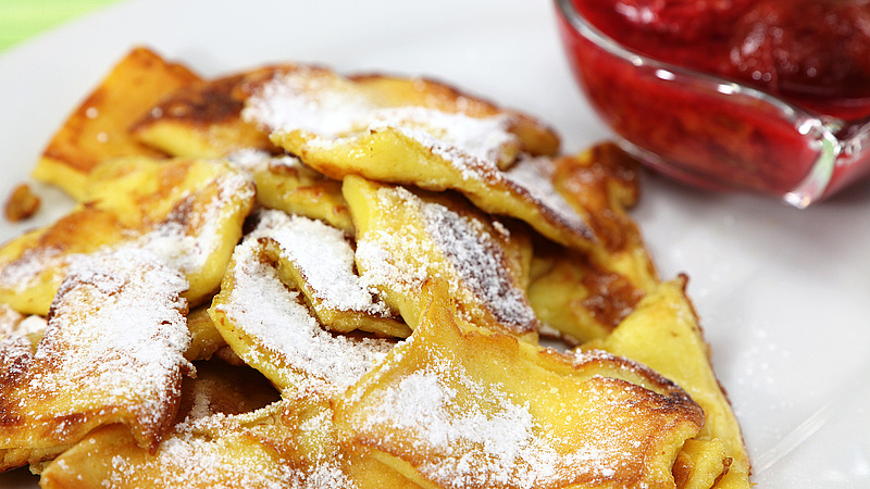 Eine Portion Kaiserschmarrn mit Puderzucker. Rechts oben im Anschnitt ein Schälchen roter Marmelade.