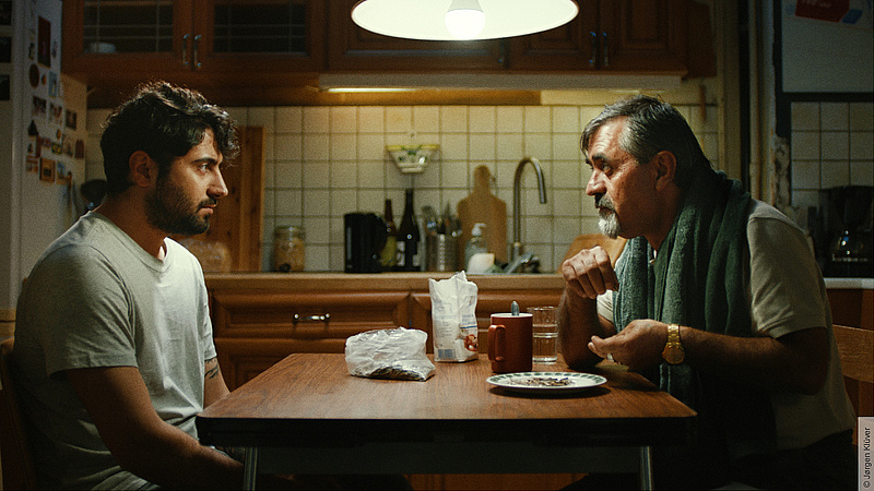 Auf dem Foto sieht man ein Foto einer Szene des Films "My Uncle Jens". Unter dem Schein der Esstischlampe sitzen zwei Männer an einem Küchentisch. Einer der beiden ist in seinen 30ern, der andere ist älter. Auf dem Tisch steht eine Tüte mit Kürbiskernen, eine offene Packung von etwas zu essen sowie eine Tasse und ein Teller. Die beiden Männer schauen sich ernst an.