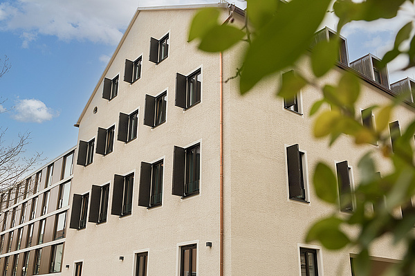 Inklusionswohnheim Kaulbachstraße - Ansicht des Altbaus Auf dem Foto sieht man im Vordergrund ein lachsfarben gestrichenes Haus mit spitzem Dach und vielen anthrazitfarbenen Fensterläden. An das Haus schließt sich ein kupferfarben verkleideter Neubau mit flachem Dach an. Im Vordergrund rechts sieht man etwas unscharf die Zweige eines Busches.