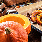 Im Vordergrund des Bildes ist ein halbierter und ausgehöhlter Hokkaido-Kürbis zu sehen. Im Hintergrund ist ein Backblech mit Backpapier erkennbar. Darauf liegen mehrere Stücke des Kürbisses, die zuvor im Ofen geröstet wurden.