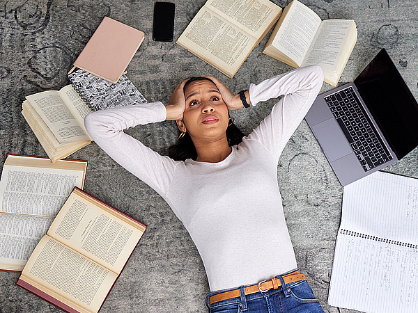 Eine verzweifelte Studentin liegt auf dem Boden umringt von geöffneten Büchern und einem Laptop. 