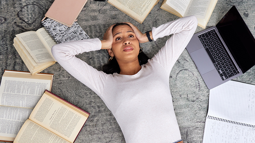 Eine verzweifelte Studentin liegt auf dem Boden umringt von geöffneten Büchern und einem Laptop. 