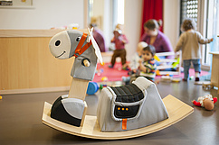 Ein gepolstertes Schaukelpferd steht im Vordergrund eines hellen Spielraums. Im Hintergrund spielen Kleinkinder auf einem Teppich, begleitet von Erwachsenen.