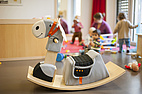 (CR) Nicolai Schneider (mit Vermerk verwendbar) Ein gepolstertes Schaukelpferd steht im Vordergrund eines hellen Spielraums. Im Hintergrund spielen Kleinkinder auf einem Teppich, begleitet von Erwachsenen.