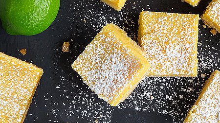 Zwei rechteckige Stücke Kokos-Limetenkuchen, die mit Puderzucker bestäubt sind, sind im Zentrum des Bildes abgebildet. Rundherum liegen weitere Stücke und eine Limette. 