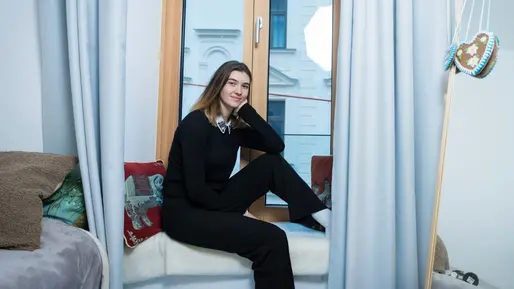 Auf dem Foto sieht man eine junge Frau, welche auf einem sehr tief liegenden Fensterbrett sitzt. Auf dem Fensterbrett liegen eine Decke und zwei Kissen. Im Vordergrund sieht man rechts neben dem Fenster Ein Bett mit Kissen im Anschnitt und links neben dem Fenster einen Spiegel, an welchem ein Lebkuchenherz hängt und vor welchem Tuben, ein Täschchen und eine Bürste liegen. Vor dem Fenster ein Teppich. Die Frau lächelt in die Kamera.