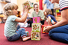 Ein blondes Kind sitzt barfuß auf einem Teppich und spielt mit bunten Stapelwürfeln. Im Hintergrund sind weitere Kinder und eine Betreuungsperson erkennbar.