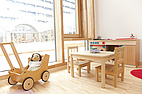 Ein heller Gruppenraum mit Kindermöbeln: Tisch mit Stühlen, Spielküche, Puppenwagen mit Puppe und Blick nach draußen durch große Fenster.