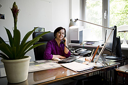 Frau in lila Bluse sitzt lächelnd an einem Schreibtisch in einem Büro und telefoniert. Auf dem Schreibtisch liegen Dokumente, eine Tastatur, mehrere Telefone und ein Computerbildschirm. Im Vordergrund steht eine grüne Topfpflanze, im Hintergrund scheint Tageslicht durch das Fenster.