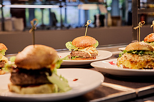 Auf dem Foto sieht man drei verschieden belegte Burger. In den Burger steckt jeweils ein kleines Spießchen. Der Burger, auf den scharf gestellt wurde, ist mit einem Fleisch-Patty und mit Salat belegt. Die Burger stehen auf Tellern auf dem Tresen einer Mensa.