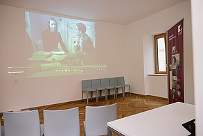 Auf dem Foto sieht man einen Raum, an dessen eine Wand ein Schwarz-Weiß-Film projiziert wird. Der Film zeigt gerade eine Frau und einen Mann, die nebeneinander an einem Tisch, auf dem viele Unterlagen liegen, sitzen. Hinter ihnen eine Tür, alles wirkt abgenutzt. In dem Raum befinden sich sonst noch einige graue Stühle, von denen manche vor der Projektion und manche an der Wand stehen. Außerdem noch ein Tisch und ein Banner, auf dem "Historie der Kaulbachstraße" steht..