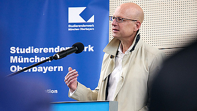 Auf dem Foto sieht man Ralph Büchele, Geschäftsführer der BayernHeim GmbH, wie er an einem Rednerpult steht und ihre Rede hält. Dahinter ein Banner mit der Aufschrift "Studierendenwerk München Oberbayern". 