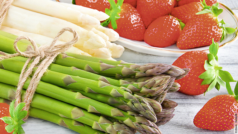 Ein Bund grüner Spargel, leicht versetzt dahinter ein Bund weißer Spargel, dahintern schöne rote Erdbeeren auf einem Teller.