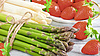 Ein Bund grüner Spargel, leicht versetzt dahinter ein Bund weißer Spargel, dahintern schöne rote Erdbeeren auf einem Teller.