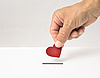 Eine Hand hält ein rotes Papierherz und wirft es in einen weißen Schlitz, der an eine Spendenbox erinnert. Das Bild symbolisiert eine liebevolle oder herzliche Spende.