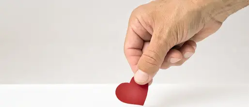 Eine Hand hält ein rotes Papierherz und wirft es in einen weißen Schlitz, der an eine Spendenbox erinnert. Das Bild symbolisiert eine liebevolle oder herzliche Spende.