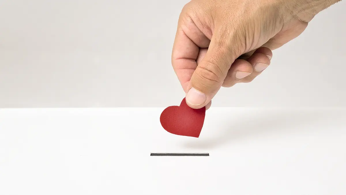 Eine Hand hält ein rotes Papierherz und wirft es in einen weißen Schlitz, der an eine Spendenbox erinnert. Das Bild symbolisiert eine liebevolle oder herzliche Spende.
