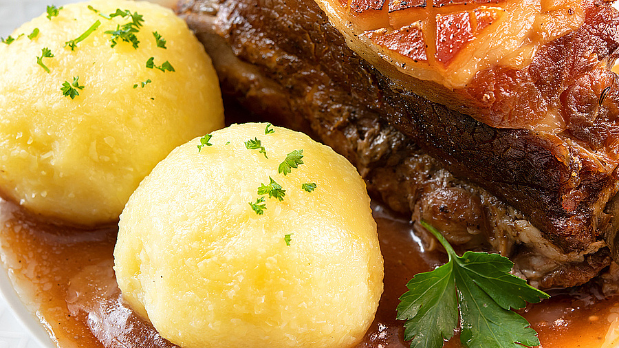 Das Bild zeigt einen Teller mit einem Stück Fleisch, zeri Kartoffelknödeln und etwas Sauce. Im Hintergrund ist eine rot-weiß-karrierte Tischdecke zu erkennen.