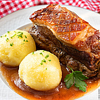 Das Bild zeigt einen Teller mit einem Stück Fleisch, zeri Kartoffelknödeln und etwas Sauce. Im Hintergrund ist eine rot-weiß-karrierte Tischdecke zu erkennen.