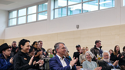 Auf dem Bild sieht man das Publikum, das klatscht. Die Personen sitzen in einer Sporthalle.