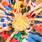 Im Zentrum des Bidles ist eine runde Pizza zu sehen. Von allen Seiten greifen verschiedene Hände nach einem Stück. Die Personen sind in unterschiedlichen Länderfllaggen gekleidet.