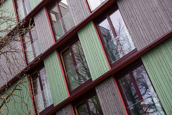 Holzfassade der Wohnanlage Schwere-Reiter-Straße Auf dem Foto sieht man die Fassade eines Hauses. Die Fenster sind schmal und bodentief. Zwischen den Fenstern erkennt man, dass die Fassade aus Holz gefertigt ist. Das Holz ist teilweise türkisfarben und teilweise anthrazitfarben angestrichen.