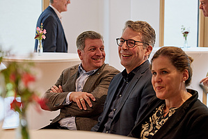 Auf dem Foto sieht man drei Personen, die nebeneinander sitzen und lachen. Es sind der Bayerische Staatsminister für Wohnen, Bau und Verkehr, Christian Bernreiter, der Bayerische Staatsminister für Wissenschaft und Kunst, Markus Blume, sowie Münchens 3. Bürgermeisterin, Verena Dietl.
