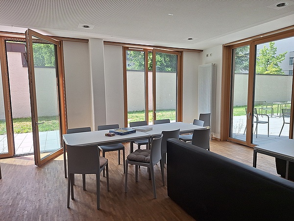 Ein Raum im Erdgeschoss mit bodentiefen Fenstern, in dem ein großer Tisch mit Plastikstühlen sowie im Anschnitt eine dunkle Couch zu sehen ist.