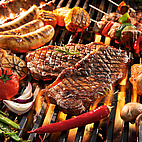 Das Bild zeigt einen Grillrost über Feuer. Darauf bruzzeln Fleisch, Würste, aber auch Gemüse.