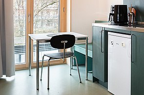 Simpler Tisch und Stuhl vor bodentiefem Fenster, rechts daneben dunkelgrüne Küchenzeile im Anschnitt