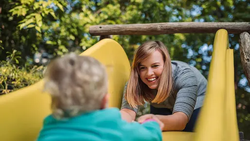 Eine lächelnde Frau hilft einem Kleinkind, die gelbe Rutsche auf einem Spielplatz hinaufzuklettern. Die Szene spielt sich im Freien bei Tageslicht ab, umgeben von grüner Natur.