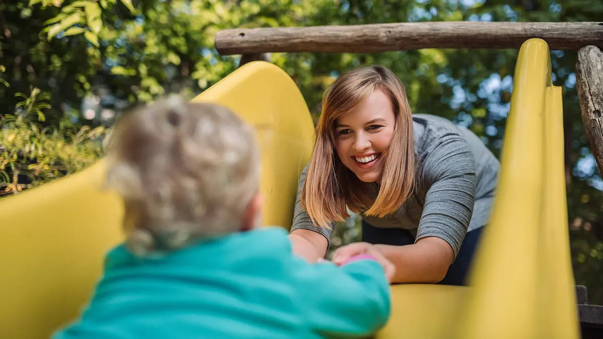 Eine lächelnde Frau hilft einem Kleinkind, die gelbe Rutsche auf einem Spielplatz hinaufzuklettern. Die Szene spielt sich im Freien bei Tageslicht ab, umgeben von grüner Natur.