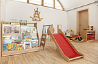 Innenraum mit Kinderbüchern in einem Regal, kleiner Rutsche, Laufrad und Spielküche. Der Raum ist hell und freundlich mit Holzfußboden eingerichtet.