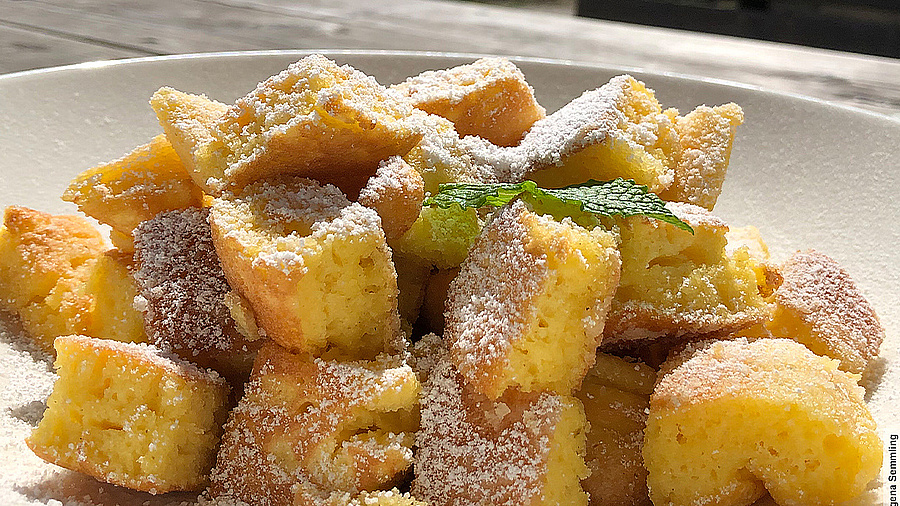 Im Vordergrund des Bildes ist ein Teller mit Kaiserschmarrn zu sehen. Dieser ist mit Puderzucker bestäubt. Im Hintergrund ist ein Alpenpanorama erkennbar.