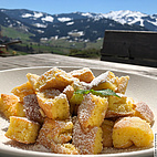 Im Vordergrund des Bildes ist ein Teller mit Kaiserschmarrn zu sehen. Dieser ist mit Puderzucker bestäubt. Im Hintergrund ist ein Alpenpanorama erkennbar.