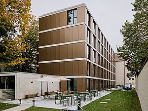 Inklusionswohnheim, Blick vom Garten auf den Neubau Auf dem Bild sieht man ein kupferfarben verkleidetes Neubaugebäude mit Flachdach von der Seite. Es steht in einem Garten mit links und rechts zwei alten Bäumen, der Baum rechts steht hinter der Mauer zum Nachbargrundstück. Im Vordergrund sieht man Wiese und eine gepflasterte Terrasse mit Gartenmöbeln..