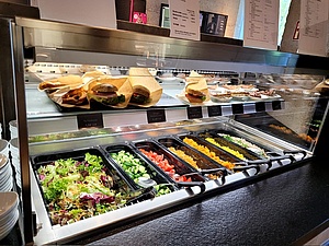 Das neu eröffnete StuBistroMensa am Bernd-Eichinger-Platz. Das Bild zeigt eine offene Essenstheke zum Selbsbedienen. Oben sind belegte Semmeln und Kuchen. Unten verschiedene Salate.