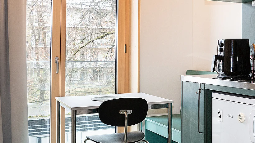 Simpler Tisch und Stuhl vor bodentiefem Fenster, rechts daneben dunkelgrüne Küchenzeile im Anschnitt
