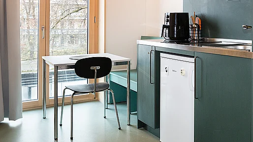 Simpler Tisch und Stuhl vor bodentiefem Fenster, rechts daneben dunkelgrüne Küchenzeile im Anschnitt