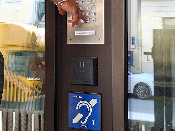 Jemand klingelt an einer Tür, unter der Klingel ein blaues Piktogramm eines Ohres, durch das ein Strich geht