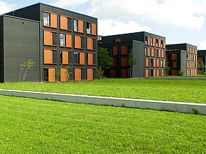 Wohnheim Stiftsbogen Auf dem Foto sieht man mehrere Neubauten und davor eine Rasenfläche. die Gebäude sind anthrazitfarben mit orangefarbenen Jalousien.